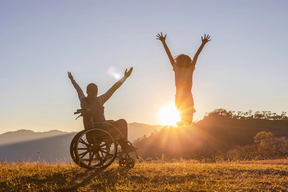Person in wheelchair with friend next to them with hands up in front of sunset